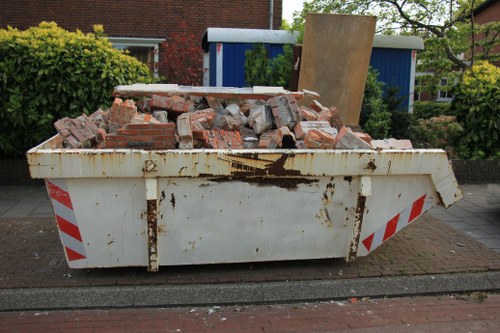 A large, white industrial skip filled with a mixture of red brick rubble and broken masonry, positioned on a street pavement in an urban area. The bricks vary in size and shape, some with rough edges and surface dirt, indicating recent demolition or clearance activity. The skip has visible rust and dirt marks on its sides, with red and white reflective stripes on the corners to enhance visibility. Behind the skip, there is a blue storage container or shed, partially obscured by a wooden board leaning against it, and lush green shrubs and small trees on either side, suggesting a residential or semi-commercial setting. The environment appears to be during daylight, with natural lighting highlighting the textures and colours of the rubble and surroundings. This scene relates to rubbish removal services provided by Garden Clearance Balham, focusing on the disposal of building and garden debris.
