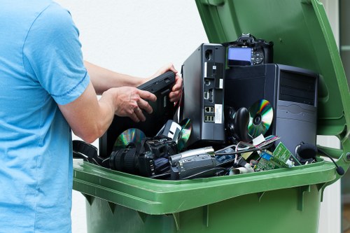 A person wearing a light blue T-shirt is seen in the process of emptying a green wheeled waste collection bin filled with electronic waste and miscellaneous debris. Visible inside the bin are discarded computer components, including an old tower-style desktop computer with a black case, a digital camera, and various cables and small electronic devices. The items appear to be in the middle of collection or disposal, with some stacked and leaning against each other. The environment appears to be outdoors on a paved surface, typical for rubbish removal services in Balham, London. The scene reflects proper handling of electronic waste, often part of a comprehensive rubbish or junk clearance service offered by Garden Clearance Balham, ensuring responsible disposal and recycling of unwanted electronic items.