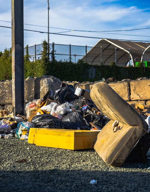 A row of black rubbish bags filled with waste, placed on a grassy area in front of a modern multi-storey residential building with large windows and a concrete facade. The bags are loosely stacked and
